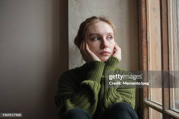 sad girl looking out window, depressed upset woman on windowsill at home. young lonely redhead woman thinking about something, pondering, people and loneliness. mood swings concept - triste foto e immagini stock