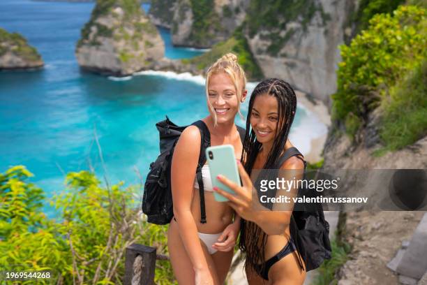 two female tourists in swimsuit taking selfie on kelingking beach on nusa penida island, bali, indonesia - animal waving stock pictures, royalty-free photos & images