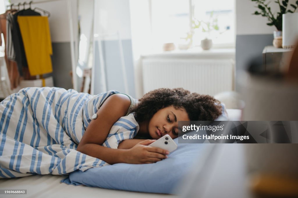 Teenage girl with diabetes feeling tired and fatigued during the day, resting in bed and scrolling on smartphone. Girl with a continuous glucose monitor on her shoulder sleeping in bed.