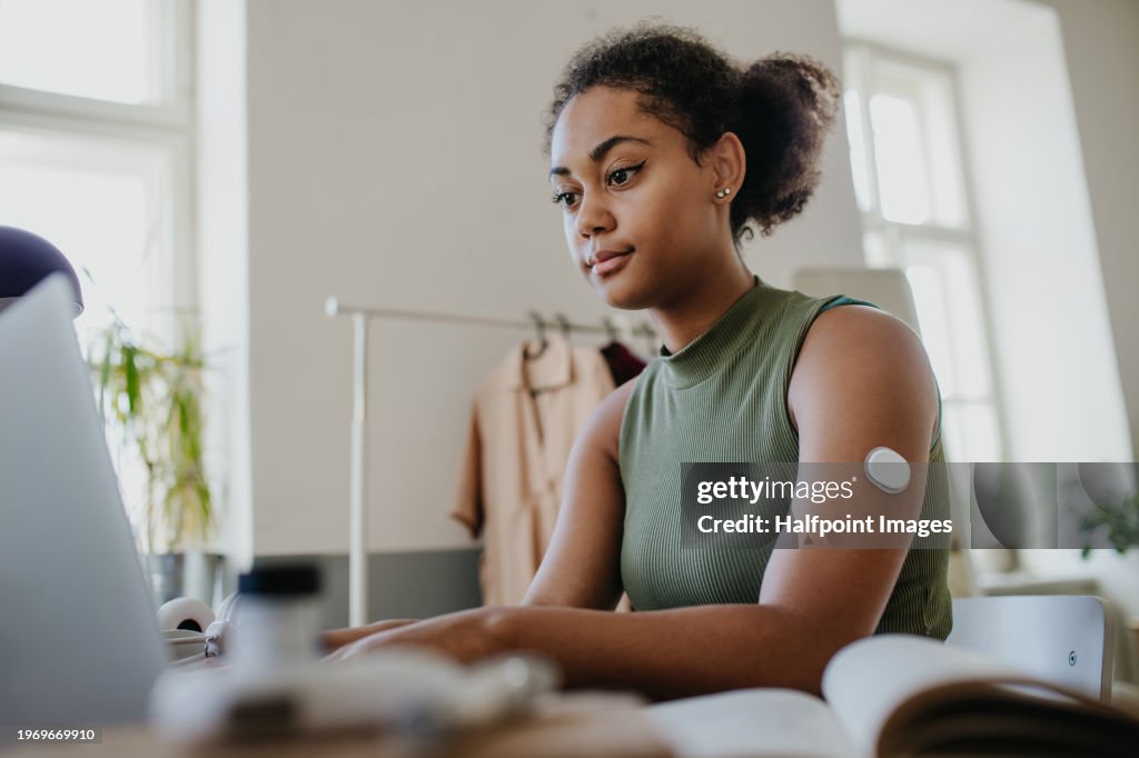 Teenage girl studying at home, doing homework while wearing continuous glucose monitor at arm.