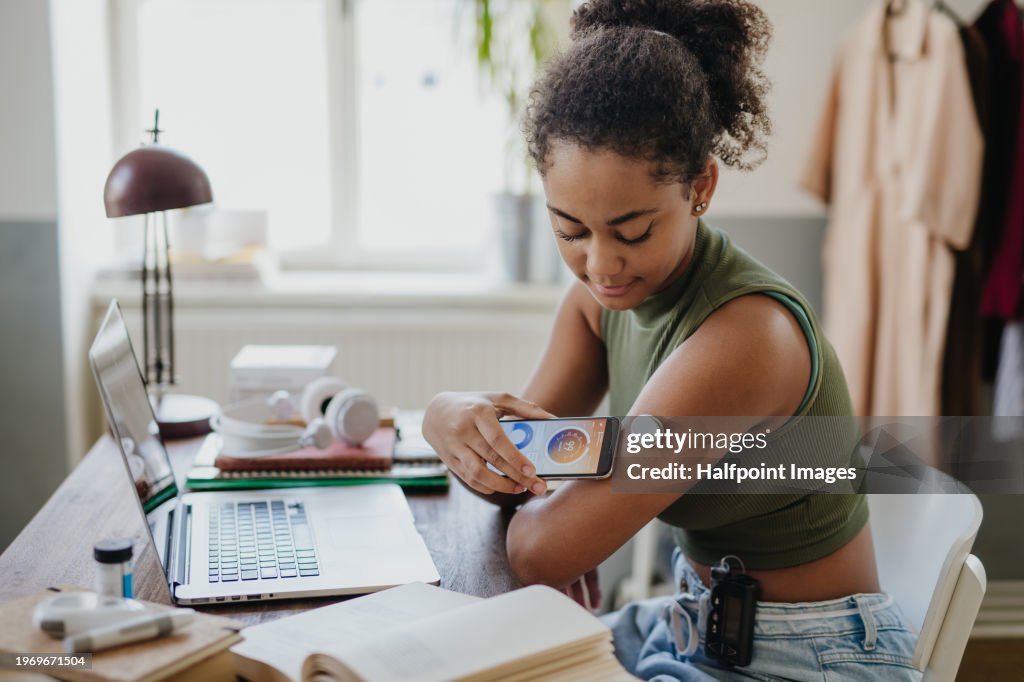 Teenage girl connecting smartphone to continuous glucose monitor, checking blood glucose during day. Student doing routine blood check while studying.