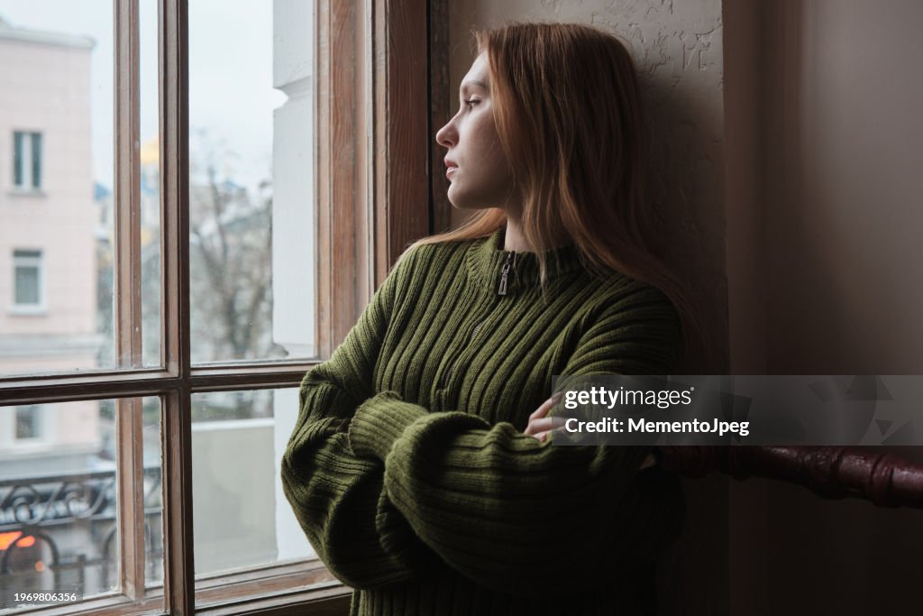 Upset lonely millennial girl standing looking out window, feeling sad and depressed. Female solitude, loneliness concept