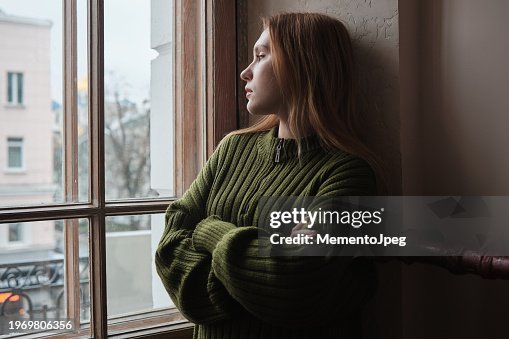 Upset lonely millennial girl standing looking out window, feeling sad and depressed. Female solitude, loneliness concept Upset lonely millennial girl standing looking out window, feeling sad and depressed. Female solitude, loneliness concept