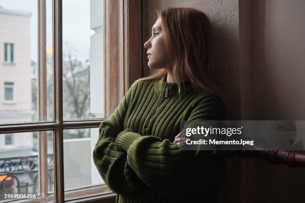 upset lonely millennial girl standing looking out window, feeling sad and depressed. female solitude, loneliness concept - abandonado imagens e fotografias de stock