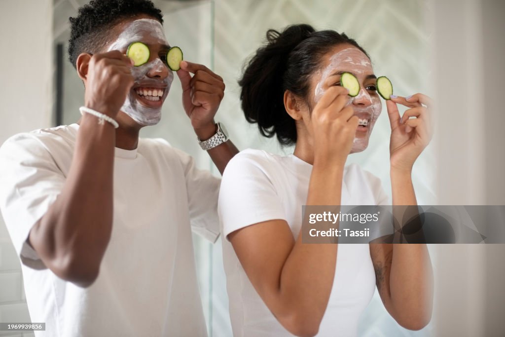 Happy couple, face and cucumber with mask for health, wellness or facial treatment together at home. Young man and woman smile with organic vegetables on eyes for anti aging, grooming or hygiene