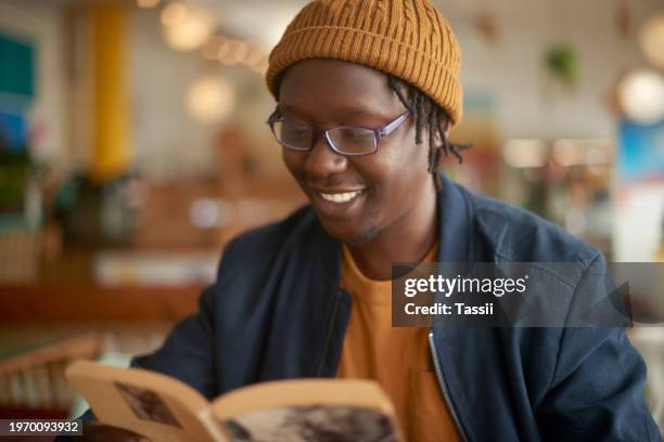 feliz, leyendo y hombre con un libro en la cafetería para la universidad, aprendiendo y estudiando el idioma en la cafetería. africanos, estudiantes y personas con investigación filosófica en el campus o disfrutar de historias de la historia - filosofía fotografías e imágenes de stock