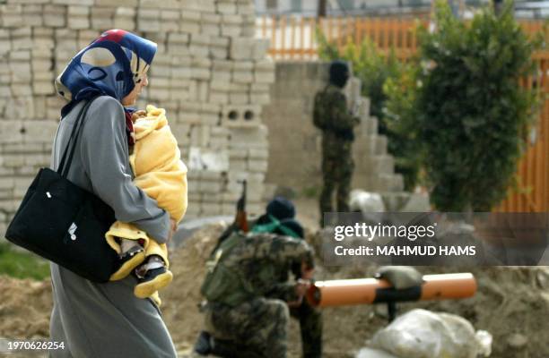 Palestinian woman carrying her child passes by militants of the radical Hamas movement preparing a makeshift anti-tank weapon during an Israeli army...