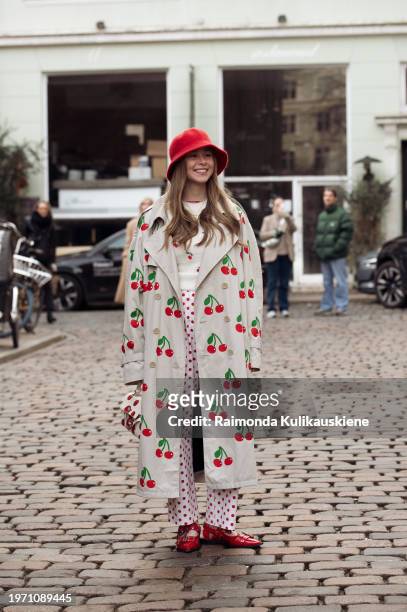 Anais Van Oekel is wearing a white top, white pants with red dots, beige coat with red cherries, red Ganni shoes, and a red wool hat during the...
