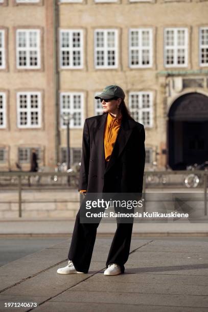 Dia Jovanovic is wearing black pants, an orange top, black jacket, white and grey New Balance sneakers, a dark green hat, and black sunglasses during...