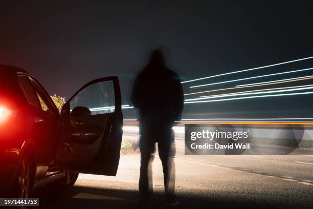 a dark mysterious blurred figure standing on a road, next to car. next to traffic trails on a country road at night - headlamp stock pictures, royalty-free photos & images