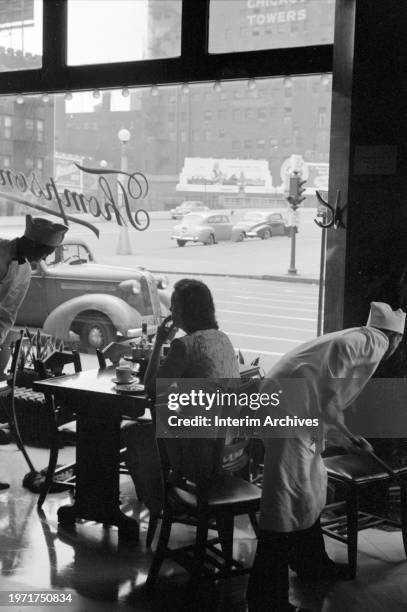 Waiter leans in as a woman sits by herself at a table in a Thompsons Restaurant cafe in Chicago, Illinois, July 1941. Another waiter mops the floor...