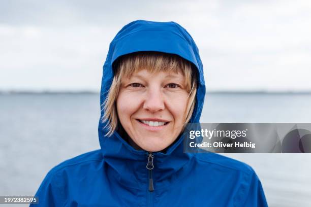 a blond woman, wearing a blue jacket with a hood, is smiling. the sea in the background is calm and clear. - regnkläder bildbanksfoton och bilder