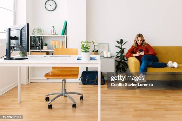 full length view of young woman using mobile phone while taking a break from work sitting on yellow sofa at modern home office. - nuria stock pictures, royalty-free photos & images