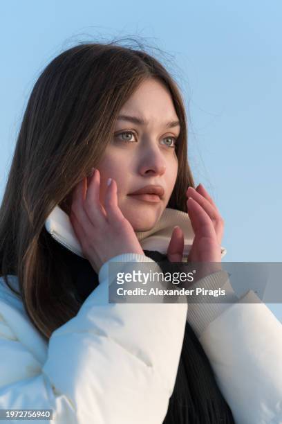 portrait of serene 17-year-old teenage girl looking away from the camera against a blue, cloudless sky on a frosty winter's day. her hands raised to face are in front of her face in a pose with her head in the center of the frame - blank frame stockfoto's en -beelden