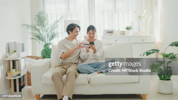 young asian couple having fun using smartphone devices shopping use credit card online payment together sit on sofa in living room at home. lifestyle spend time in house. - online bank statement stock pictures, royalty-free photos & images