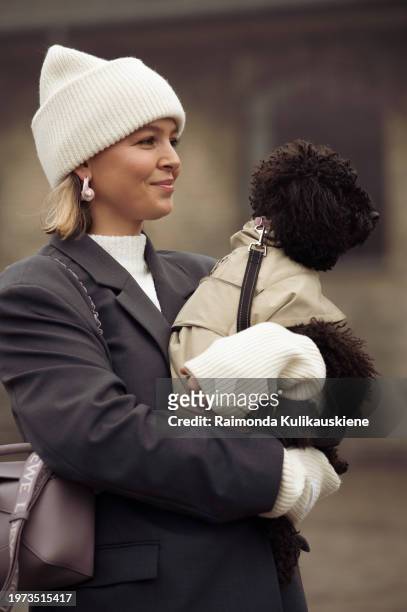 Guest with Toy Puddle dog wears a grey suit, white hat, white sweater, light pale purple Loewe bag, and white Ganni shoes outside Rolf Ekroth during...