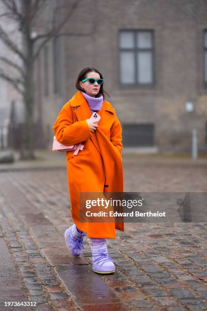 Guest wears green sunglasses, a purple oversized ribbed pullover / long dress, neon orange long coat, purple moon boots, outside Rolf Ekroth, during...