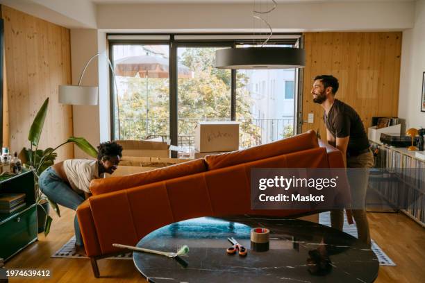 multiracial couple helping each other while lifting sofa in living room at new home - körperliche aktivität stock-fotos und bilder