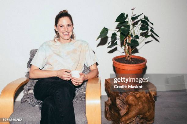 portrait of smiling woman holding tea cup while sitting on chair by plant at home - flash stock pictures, royalty-free photos & images