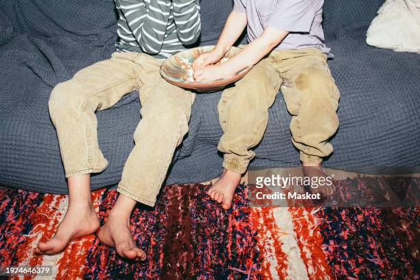 low section of male siblings sitting with popcorn bowl on sofa at home - front flash photography foto e immagini stock