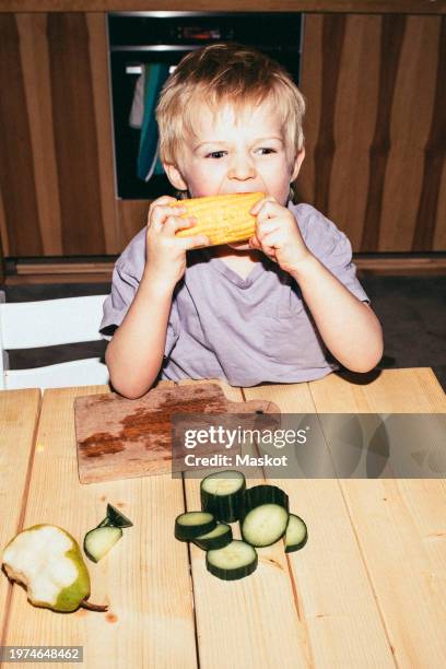 hungry boy biting corn while sitting at table in home - flash stock pictures, royalty-free photos & images