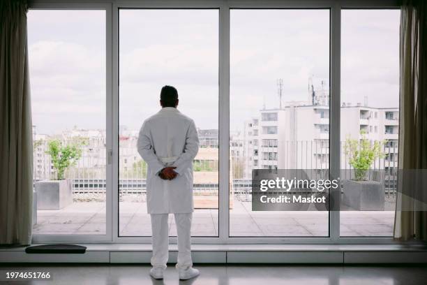full length rear view of doctor with hands behind back looking out through window at hospital - mãos-atrás-das-costas imagens e fotografias de stock