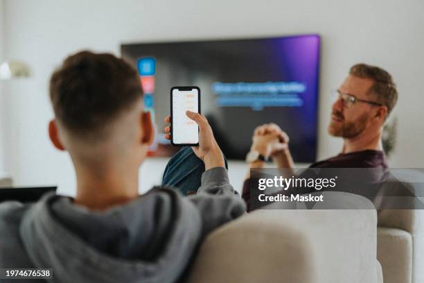 teenage boy with mobile phone talking to father while sitting in living room - smart tv fotografías e imágenes de stock