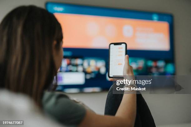 girl using smart phone app while watching tv in living room at home - smart tv stock-fotos und bilder