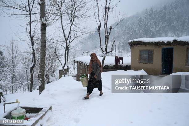 Woman walks to fetch water from a tap outside her mud house during a snowfall on the outskirts of Srinagar on February 3, 2024.