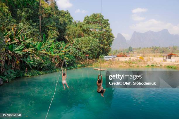 man and woman enjoying a rope swing over a serene blue lagoon in a tropical setting - vang vieng stock pictures, royalty-free photos & images