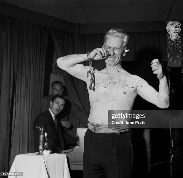 British circus performer Eddie Sinclair pushes a nail through his nose, hatpins, pins and needles pinned to his bare chest, and a pair of handcuffs...