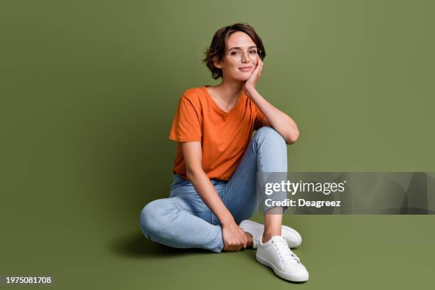 Full length photo of positive cheerful girl wear stylish clothes sit on floor hand on cheekbone isolated on green color background