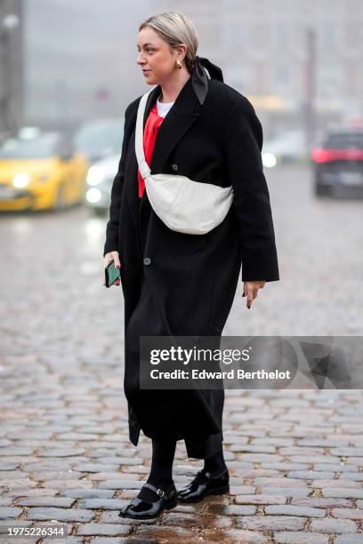 Guest wears golden earrings, a black long coat, a white leather fanny pack bag, black leather shiny shoes, outside Rolf Ekroth, during the Copenhagen...