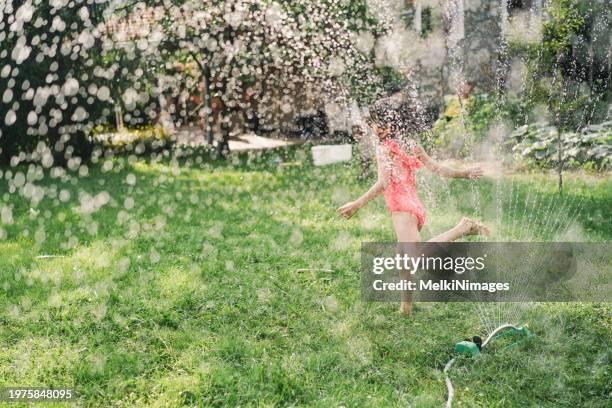 little girl having fun jumping over garden sprinkler and refresh herselves on a hot summer day - sprinkler stock pictures, royalty-free photos & images