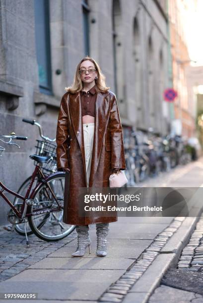 Guest wears sunglasses, a brown shiny leather long trench coat, a brown cropped shirt, a white skirt, high heels gathered boots , outside Herskind,...