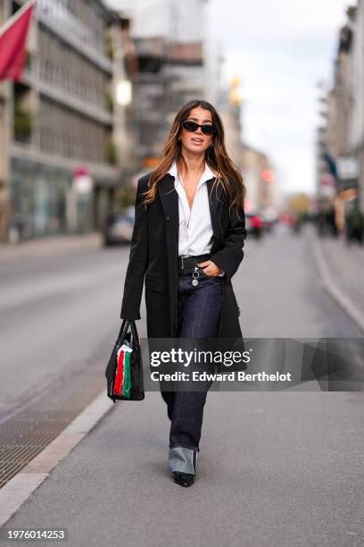 Guest wears sunglasses, a white shirt, a black long oversized blazer jacket , a belt, blue flared denim jeans pants , a Hermes bag, pointed shoes,...