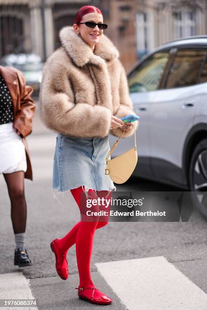 Marianne Theodorsen wears sunglasses, a beige oversized fluffy faux fur jacket , blue ripped mini shorts, red leggings, red flat shoes, a beige bag,...