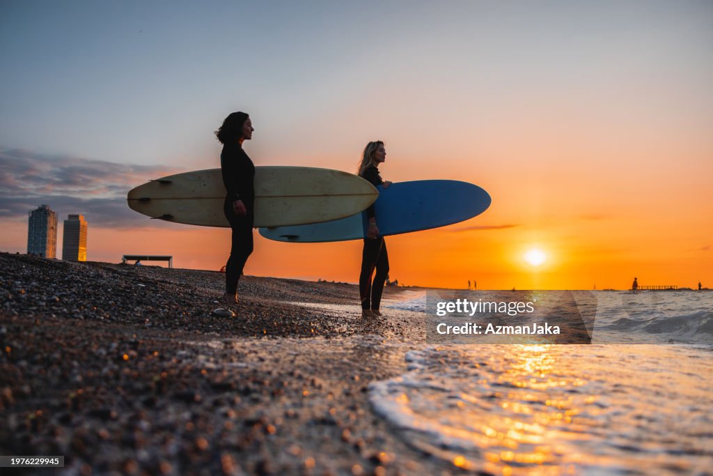 Two Female Surfers Stopping By The Water To Admire The Amazing View Before A Surf Session At Sunrise