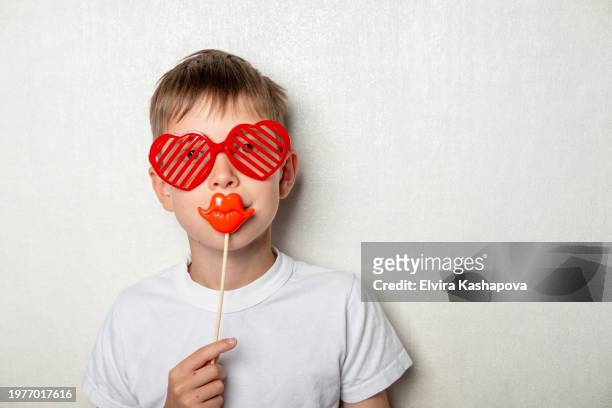 portrait of a boy 10 years old in a white t-shirt and heart-shaped glasses and a mask with lips on a white wall - 10 11 years stock pictures, royalty-free photos & images