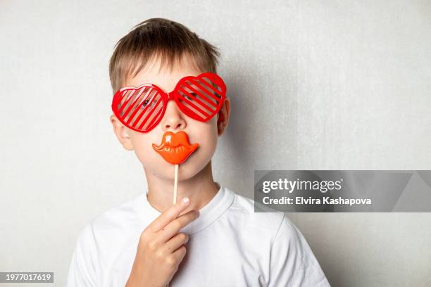 portrait of a boy 10 years old in a white t-shirt and heart-shaped glasses and a mask with lips on a white wall - 10 11 years stock pictures, royalty-free photos & images