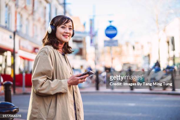 cheerful asian businesswoman listening to music over wireless headphones from smart phone while commuting in the city - charming stock pictures, royalty-free photos & images