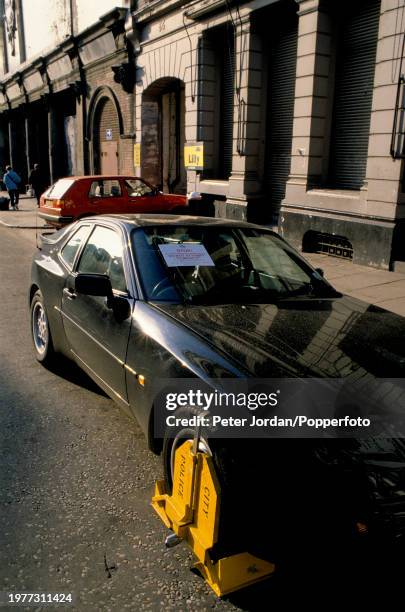 Wheel clamp fitted by a parking enforcement team to the front wheel of a Porsche 944 sports car parked on a street in the Smithfield Market district...