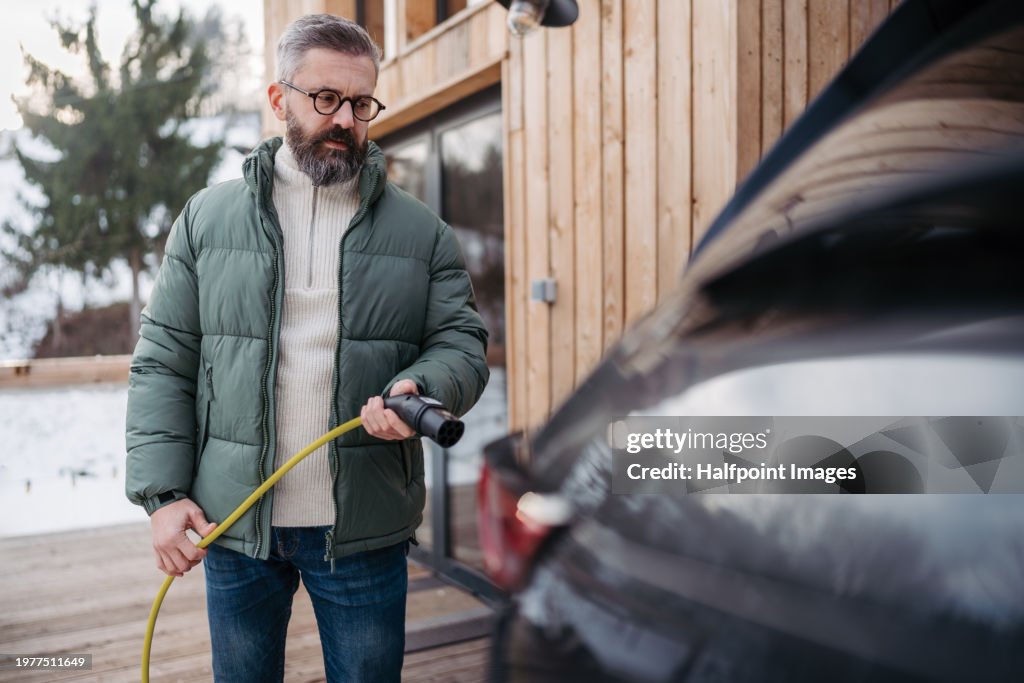Man charging electric car in front his cabin in mountains. Electric vehicle with charger in charging port. Man driving electric car on winter holiday in mountains.