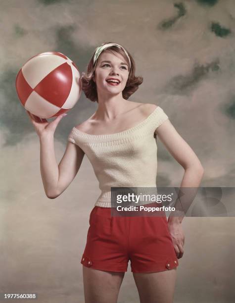 Posed studio portrait of a female fashion model wearing a pair of red shorts and white knitted top with scoop neck, she holds a beach ball in one...