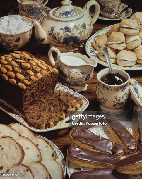 Rich fruit cake, scones with jam and cream, chocolate eclairs and slices of fruit bread and butter, all laid out on plates on a wooden table for...