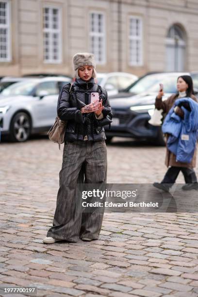 Guest wears a gray fluffy chapka hat, a black puffer jacket, flared pants , beige shoes, holds a mobile phone to take a picture / selfie, outside...