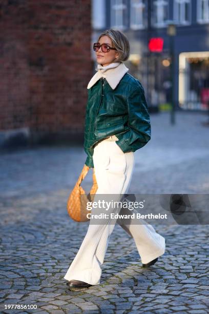 Guest wears sunglasses, a green leather jacket with white fluffy collar, white flared pants palazzo pants , a brown woven leather bag, outside...