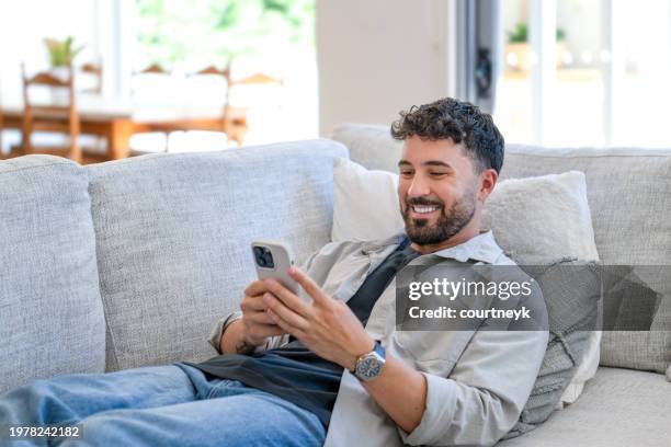 joven feliz que se relaja sentado y reclinado en el sofá de casa. está usando un teléfono móvil. - hombres-jóvenes fotografías e imágenes de stock