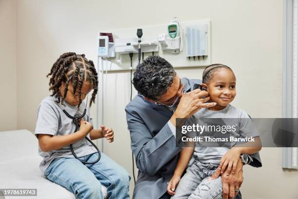 medium shot doctor looking in young patients ears in medical exam room - ear exam stock-fotos und bilder