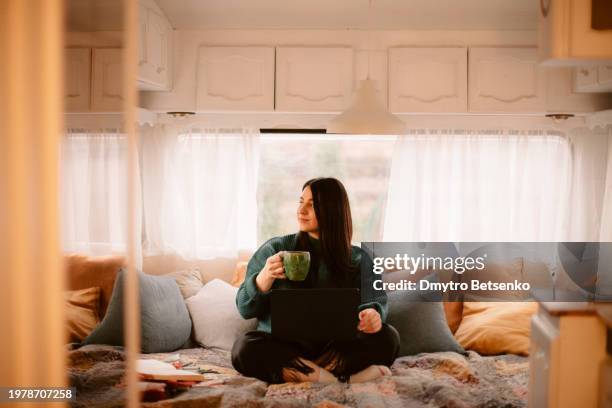 young woman holding mug with hot drink while using laptop computer sitting on the bed in the camper trailer - caravan interior stock pictures, royalty-free photos & images
